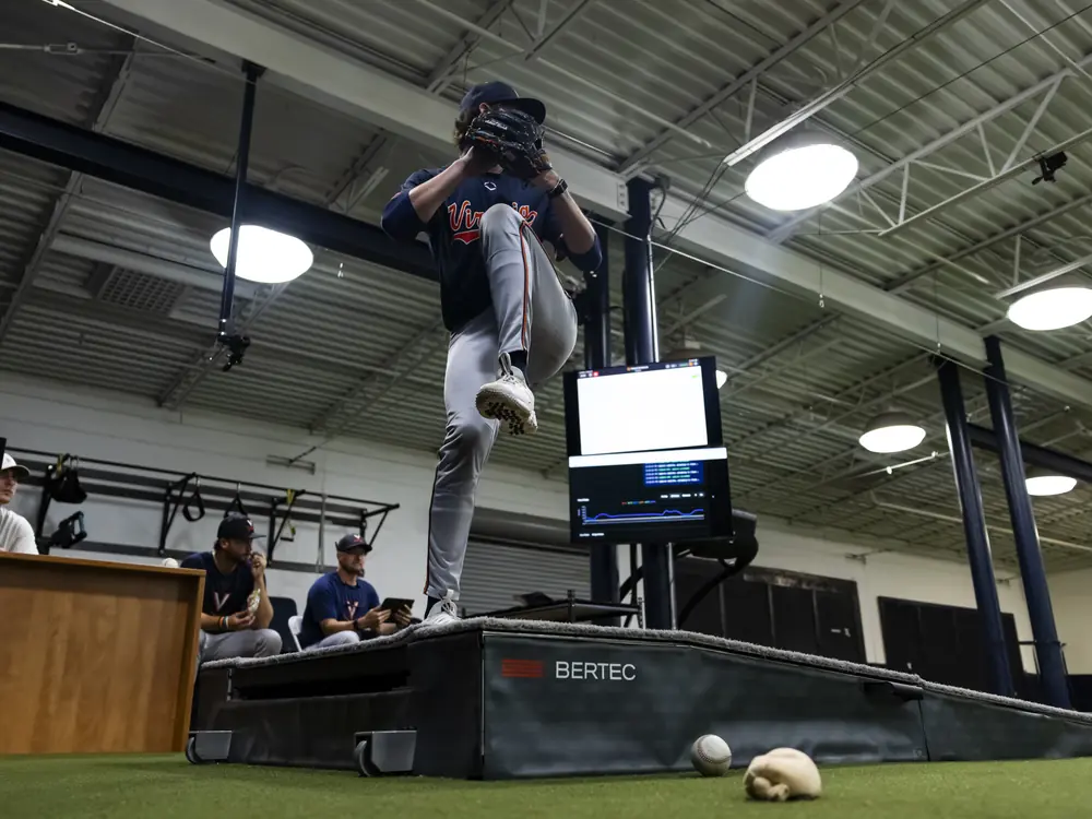 A Virginia pitcher mid-delivery on the lab mound while coaches watch from a desk behind him